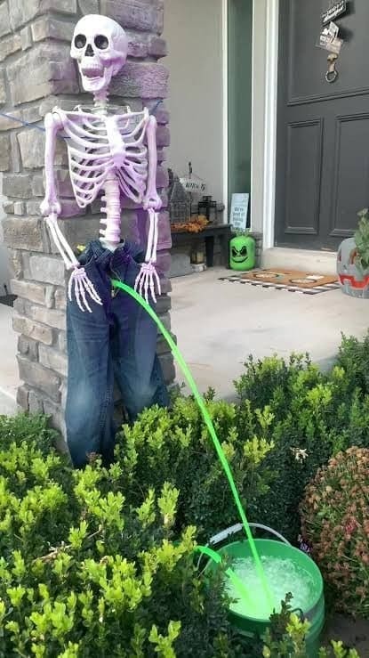 Skeleton decoration in jeans with a green hose streaming into a bucket by the porch.