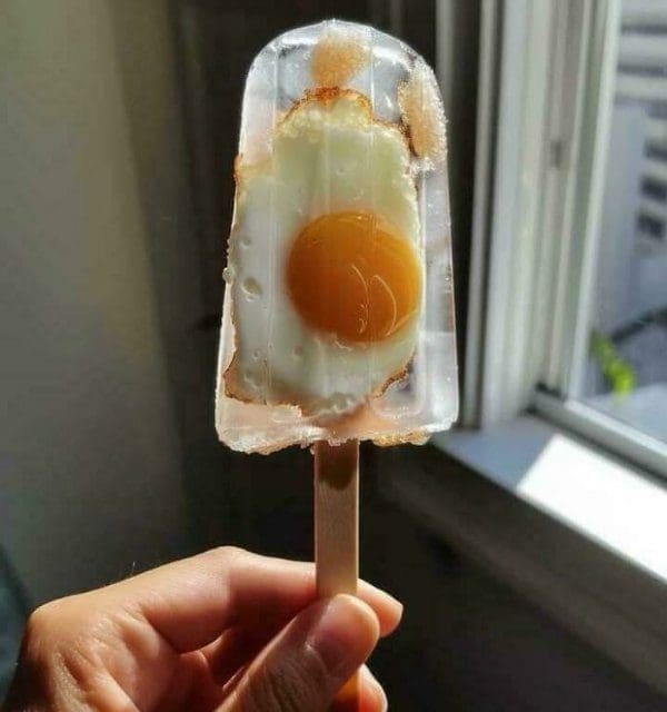 Hand holding a clear popsicle with a whole fried egg frozen inside it.