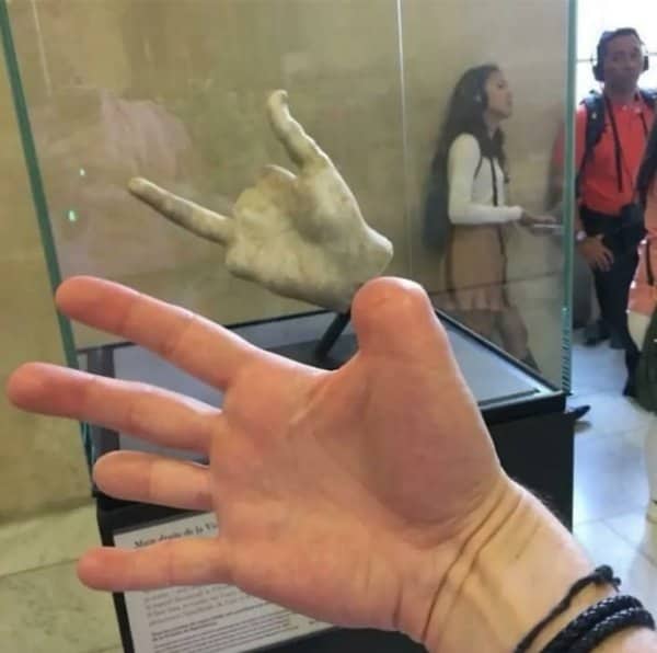 Tourist holding up an open hand beside a broken ancient marble hand in a museum display case.