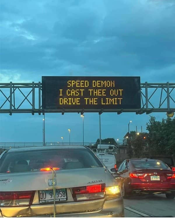 Highway message board reading Speed Demon I Cast Thee Out, Drive the Limit, above evening traffic.