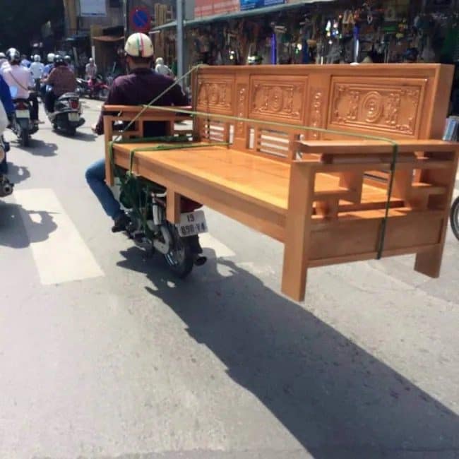 Meme dump opener of a moped rider hauling a full wooden bench through city traffic