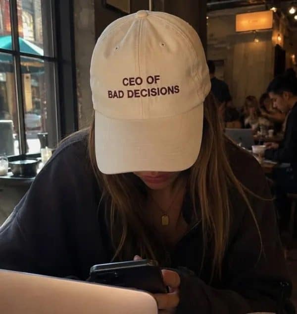 Woman in a cafe looking at her phone while wearing a CEO of Bad Decisions cap