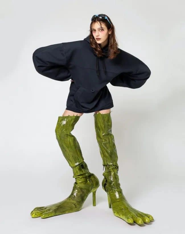 Model in an oversized black hoodie wearing glossy green thigh boots shaped like giant feet.