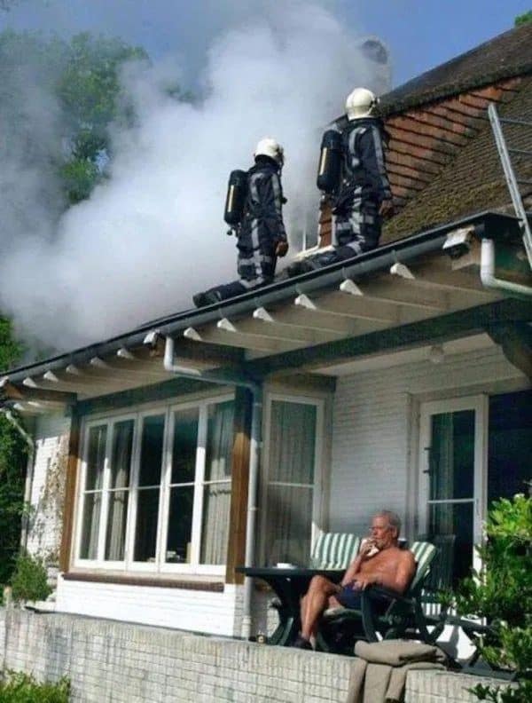 Shirtless man smokes calmly on his patio while firefighters work on his smoking roof above.