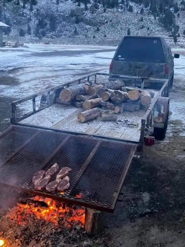 Steaks grilling over a fire on a flatbed trailer loaded with firewood in the snow.