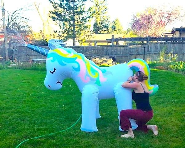Woman kneeling behind a giant inflatable unicorn sprinkler in a backyard, an odd meme dump photo.