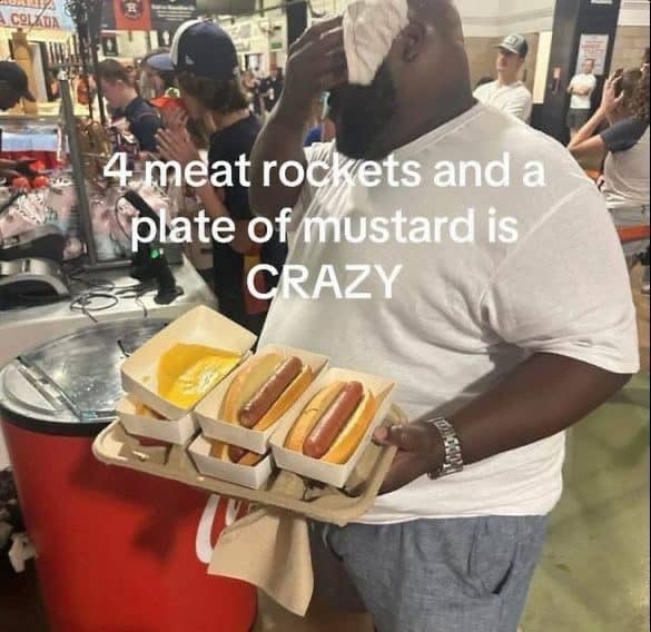 Man at a ballpark concession stand carrying a tray of four hot dogs and a cup of mustard.