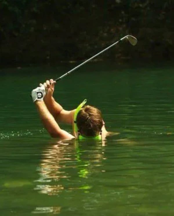 Golfer standing chest deep in a lake, still swinging a club to hit the ball.
