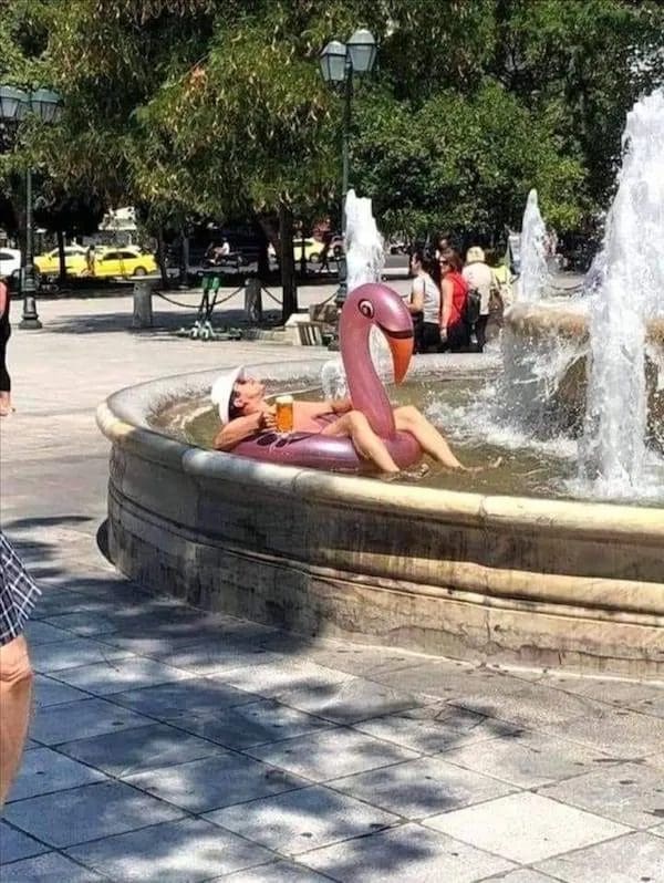 Man relaxing on an inflatable flamingo with a beer inside a public city fountain.
