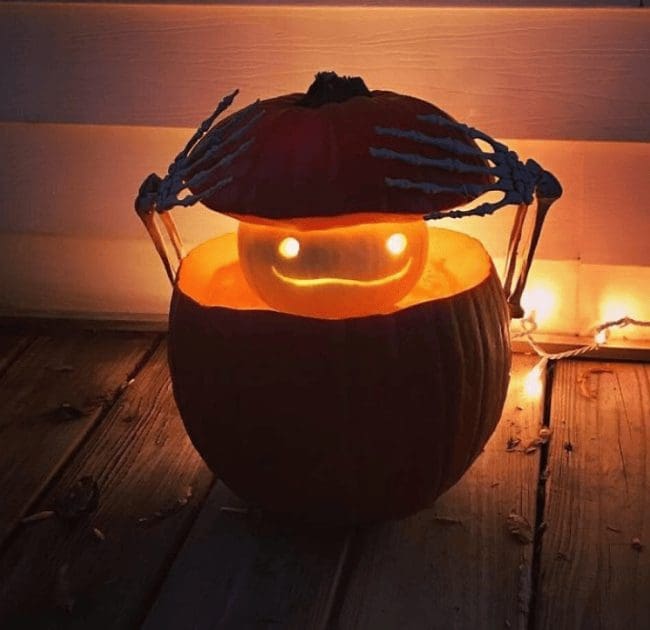 Skeleton hands lifting the lid of a pumpkin to reveal a smaller glowing pumpkin inside.