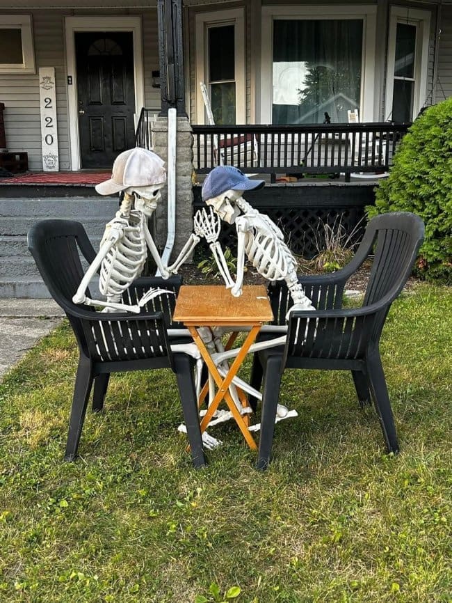 Two skeletons in ball caps arm wrestling across a small table between lawn chairs.