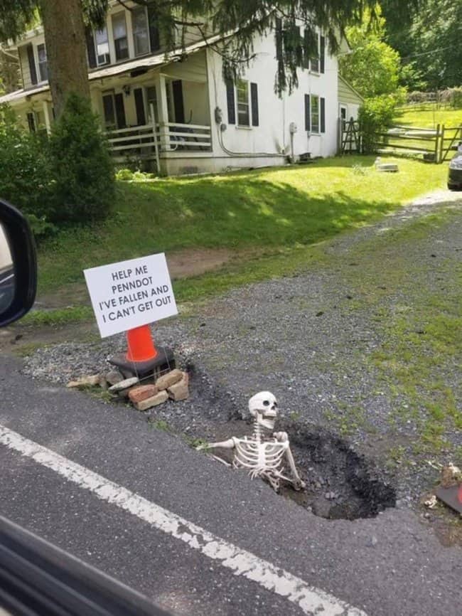 Skeleton stuck in a roadside pothole with a sign asking the state transportation department for help.