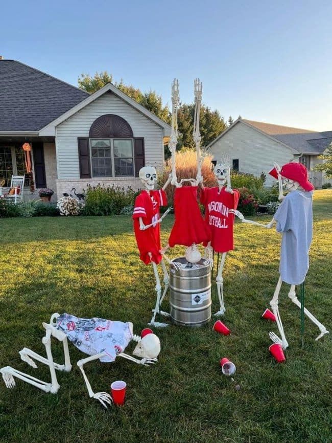 Skeletons in college football jerseys doing a keg stand with red cups scattered on the lawn.