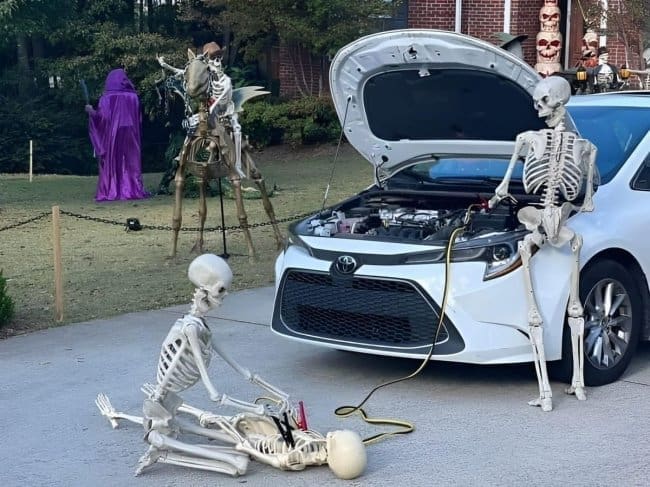 Halloween skeletons jump starting a car in a driveway while another lies collapsed beside them.