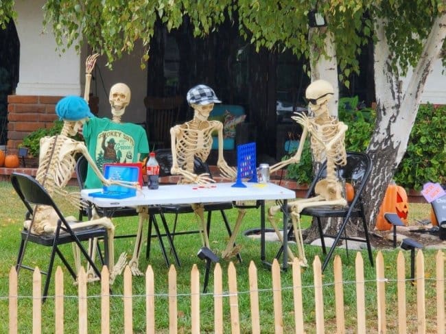Four skeletons in hats playing Connect Four and drinking soda at a folding table.