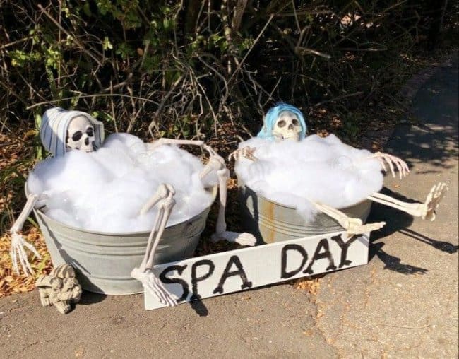 Two skeletons relaxing in metal tubs filled with bubbles behind a spa day sign.