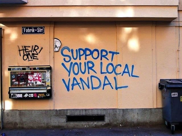 Blue graffiti on a peach building wall reading support your local vandal.