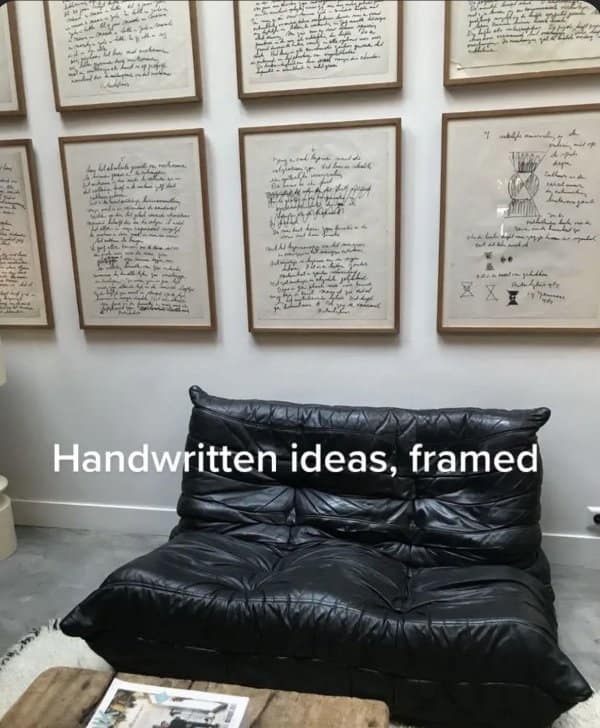 Framed handwritten pages covering a wall above a black leather lounge sofa on a gray floor.