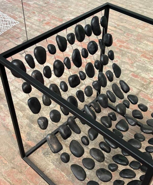 A black metal cube frame strung with dozens of smooth black stones hanging on thin wires.