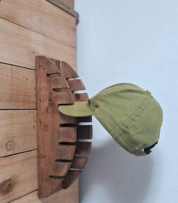A wooden wall-mounted hat rack with slotted fins and a green baseball cap clipped onto it.