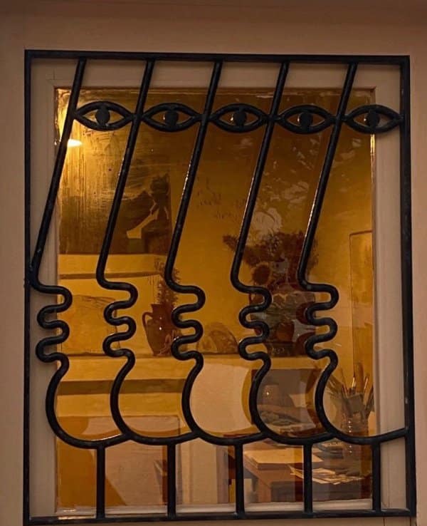 Wrought iron window bars bent into a row of side profile faces with eyes and lips.