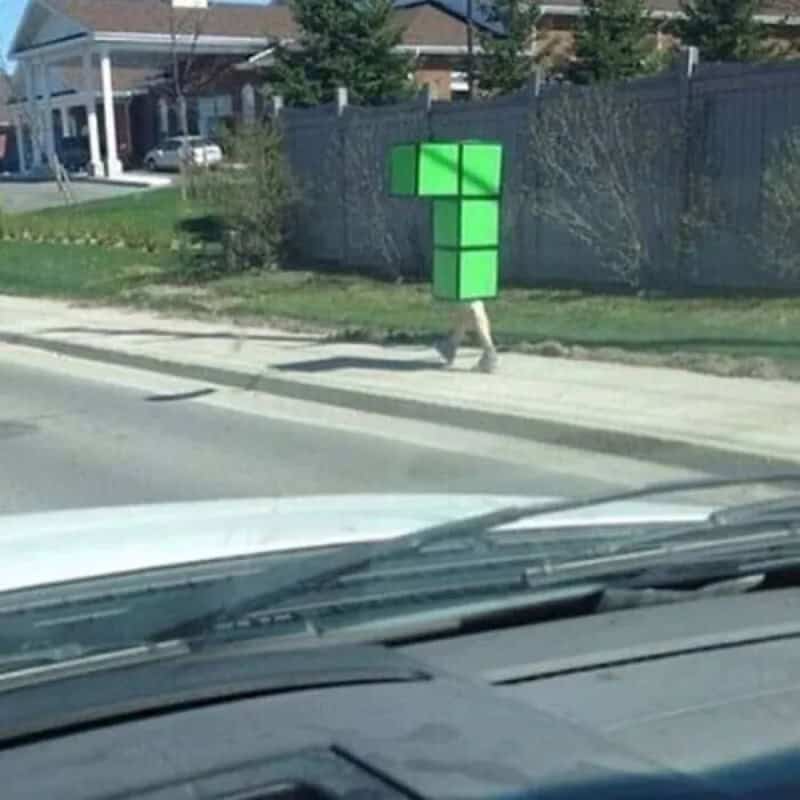 Person in a green Tetris block costume walking along a suburban sidewalk.