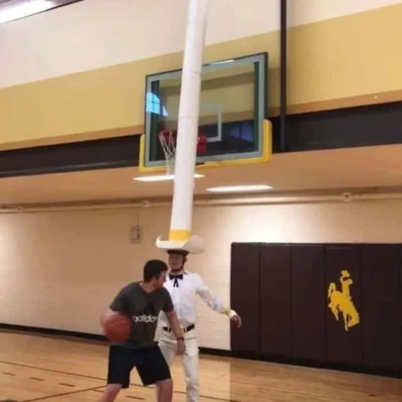 Person in a giant towering cowboy hat guarding a basketball player in a gym.