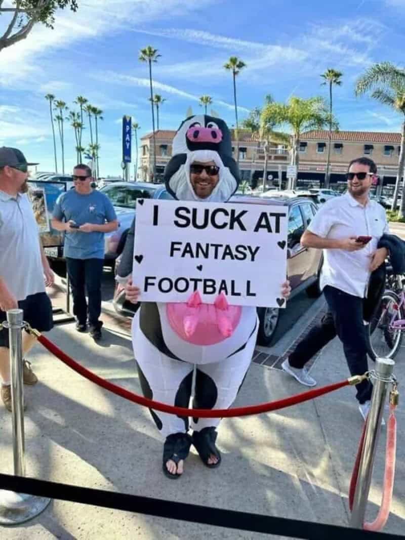 Man in an inflatable cow costume holding a sign reading I suck at fantasy football.