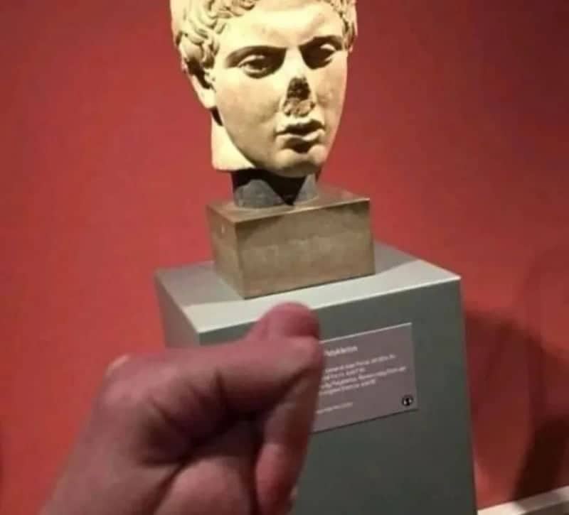Thumb held up in front of an ancient bust so it fills in the statue's missing nose.