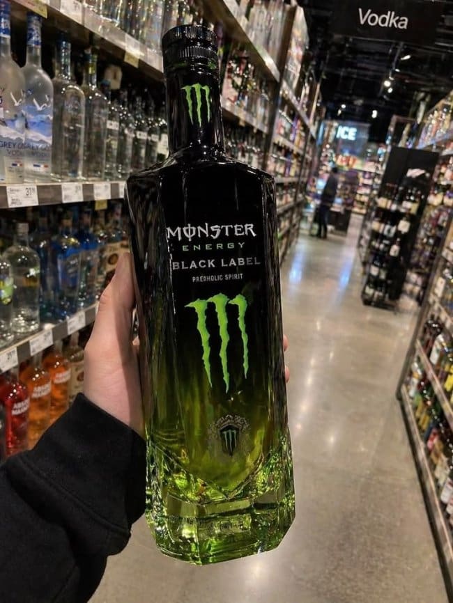 Hand holding a green and black Monster Energy liquor bottle in a store's vodka aisle.