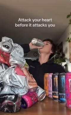 Person chugging an energy drink beside a mountain of crushed cans, joking about heart attacks.
