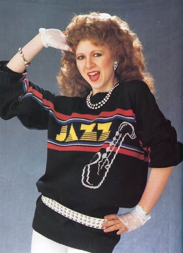 A woman in a black sweater with the word “JAZZ” and a saxophone graphic, styled with 80s jewelry and white gloves.