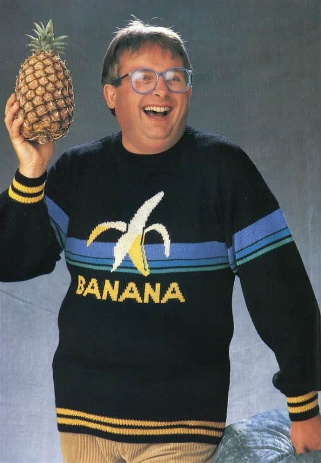 A laughing man wearing a sweater with a banana graphic and holding up a pineapple.