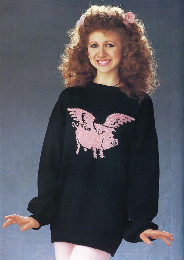 A woman in a black sweater with a pink flying pig graphic and big 80s hair tied with pink bows.