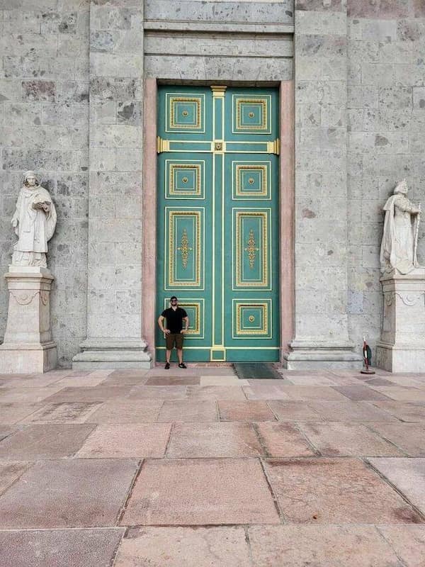 A man stands in front of a colossal green and gold cathedral door.