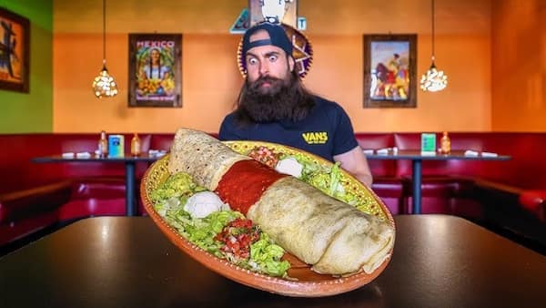 A massive burrito plated with generous servings of guacamole and salsa, dwarfing the man behind it.