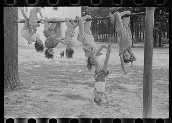 A vintage photo from a dangerous playground of many young children hanging upside down from a single metal bar.