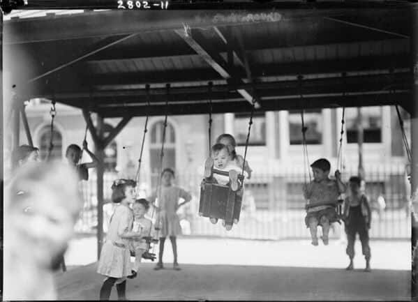 A vintage photo from a dangerous playground showing toddlers in unsafe, box-like swings with no safety harnesses.