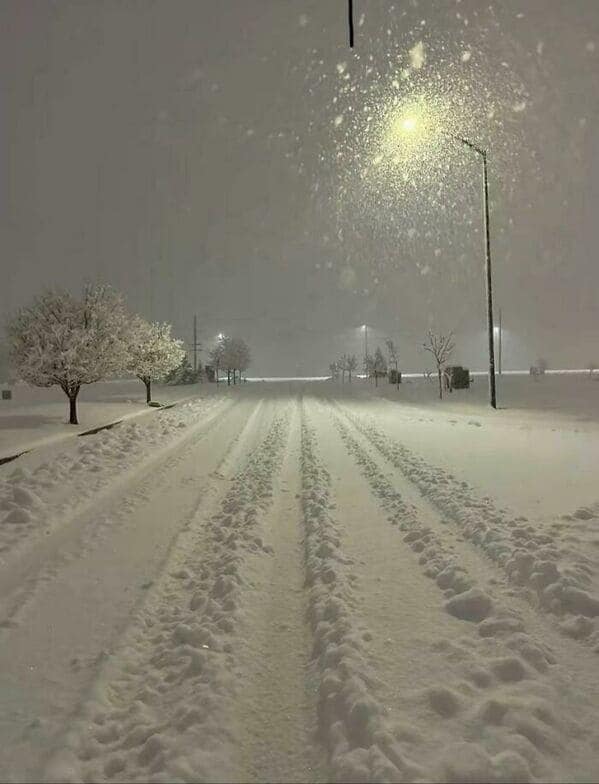 A liminal space creepy snowy road at night illuminated by a single yellow street light.