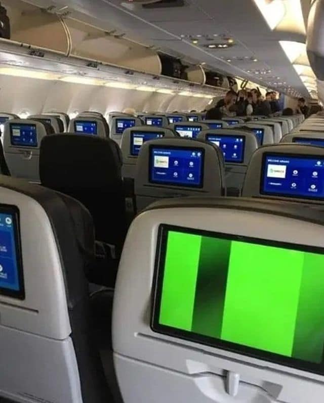A meme dump photo of an airplane cabin where all screens are green except one working screen.