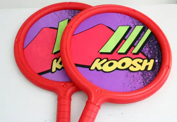 Two red plastic paddles with purple centers featuring the Koosh logo and green graphics