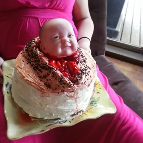 A funny cake designed for a baby shower featuring a baby doll head bursting out of the top surrounded by red strawberry filling like a horror movie scene.
