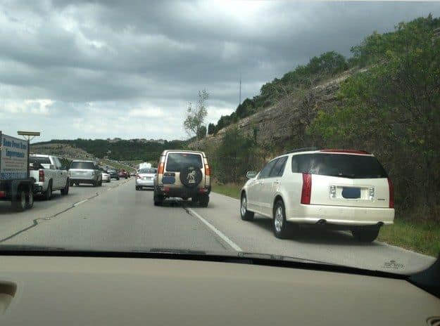 A driving meme photo of heavy traffic where a white Cadillac SUV is driving on the grass shoulder to cut the line.