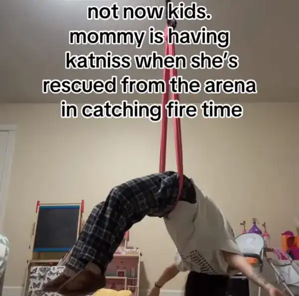 An absolutely wild unhinged screenshot of a woman suspended from the ceiling by pink silk yoga straps, arched into a backbend over a child's playroom. The caption reads: "not now kids. mommy is having katniss when she’s rescued from the arena in catching fire time."