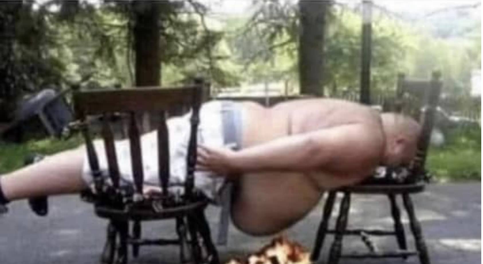 Large shirtless man balancing horizontally between two wooden chairs over a small campfire outdoor.