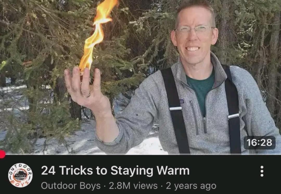 classic meme screenshot of an "Outdoor Boys" YouTube thumbnail showing a man smiling while holding a literal, flickering flame in his bare hand, titled "24 Tricks to Staying Warm."