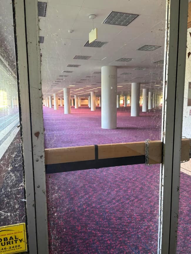 A vast, abandoned office floor with rows of repeating white support columns and pink speckled carpet is viewed through a chained glass door, embodying the architectural dread of eerie liminal spaces.