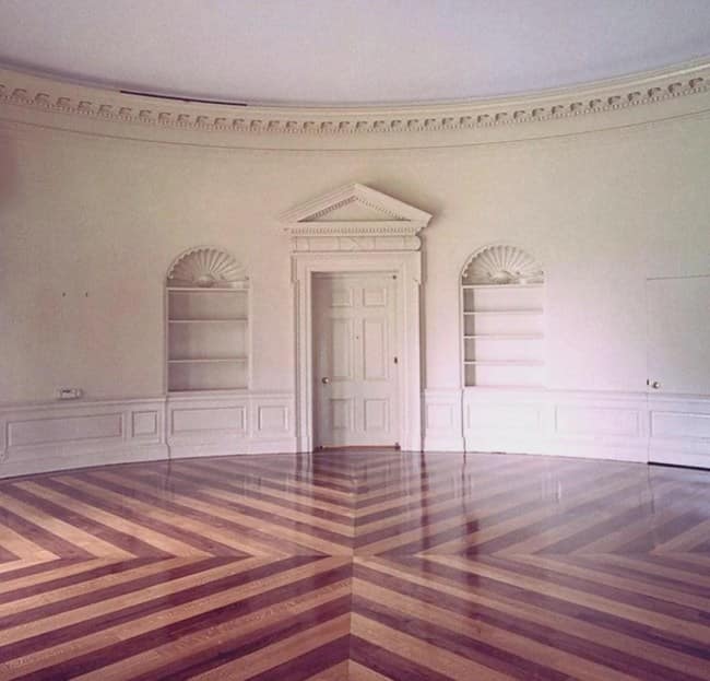 An empty, curved white room resembling a vacated Oval Office features a polished chevron-pattern wood floor and a classical pediment doorway, illustrating the quiet vacuum of eerie liminal spaces.