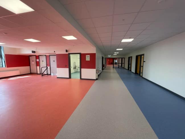 A deserted modern school corridor cleanly divided by bold red, grey, and blue floor sections under sterile ceiling tiles channels the unsettling aesthetic of eerie liminal spaces.
