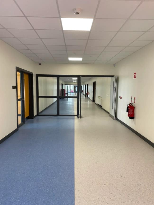 An institutional hallway floor split directly down the middle with blue and white vinyl tile leads toward a dark glass partition wall inside eerie liminal spaces.
